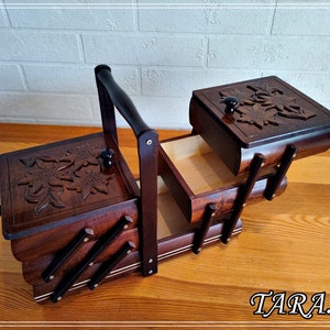 May include: A wooden sewing box with a hinged lid and a carved floral design. The box has a handle and opens to reveal two compartments. The box is made of dark wood and has a light wood interior.