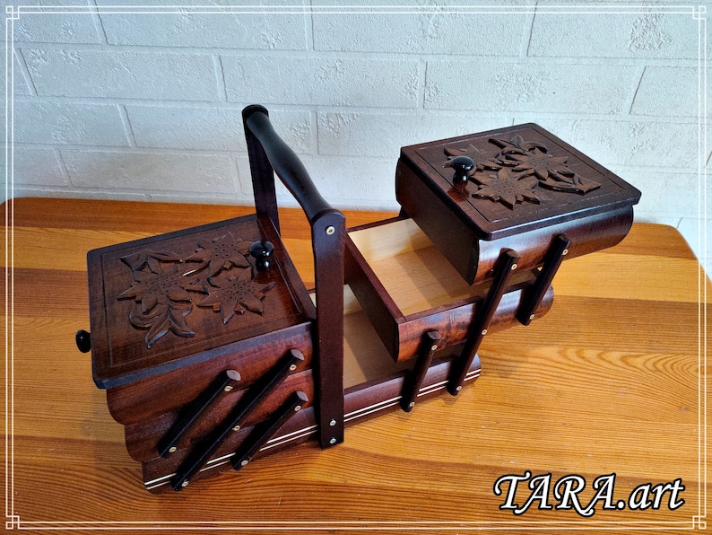 May include: A wooden sewing box with a hinged lid and a carved floral design. The box has a handle and opens to reveal two compartments. The box is made of dark wood and has a light wood interior.