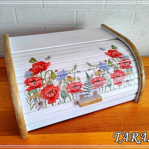 Puede incluir: Caja de pan de madera blanca con tapa enrollable. La tapa presenta un diseño floral con amapolas rojas y otras flores silvestres. La caja de pan está sobre una superficie de madera.