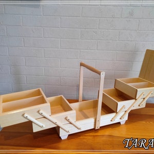 May include: A wooden sewing box with four compartments and a handle. The box is open and the compartments are visible. The box is made of light wood and has a natural finish.