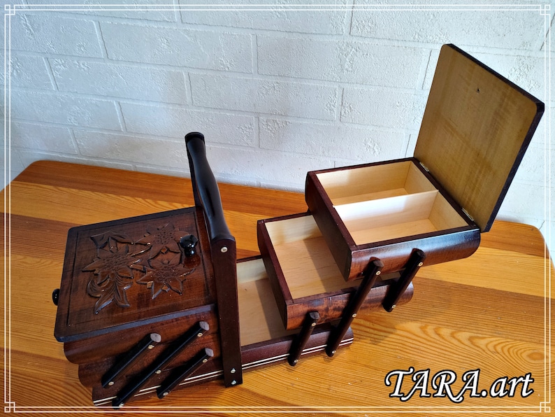 May include: A wooden sewing box with three drawers and a carved floral design on the top. The box has a handle and is open to reveal the empty drawers.