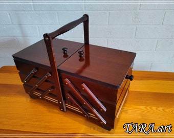 May include: A dark brown wooden sewing box with a glossy finish, featuring a cantilevered accordion-style design with multiple tiers, a central carrying handle, two top lids with small round knobs, and the text TARA.art in the bottom corner.
