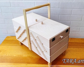 May include: A light-coloured wooden sewing box with a handle, two small knobs on the lid, and three drawers. The box has a natural wood finish and is set on small, rounded legs. The handle is also made of wood.