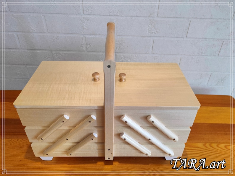 May include: A wooden sewing box with a hinged lid and a handle. The box has multiple compartments and is decorated with white wooden dowels.