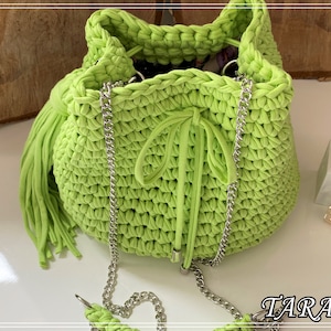 May include: A bright green crocheted bucket bag with a silver chain strap and a long green tassel. The bag has a drawstring closure and is made with a chunky yarn.