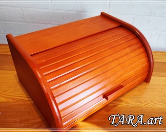 May include: A wooden bread bin with a roll-top lid. The lid is made of slats and has a small handle. The bin is a light brown colour.