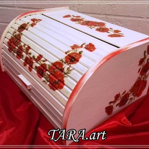 May include: White wooden bread bin with a roll-top lid. The lid is decorated with red poppy flowers and green leaves. The bin is sitting on a red fabric surface. The text "TARA.art" is printed on the bottom of the bin.