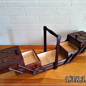 May include: A wooden sewing box with a hinged lid and a handle. The box has three compartments and is decorated with carved floral designs. The box is open and the compartments are empty.