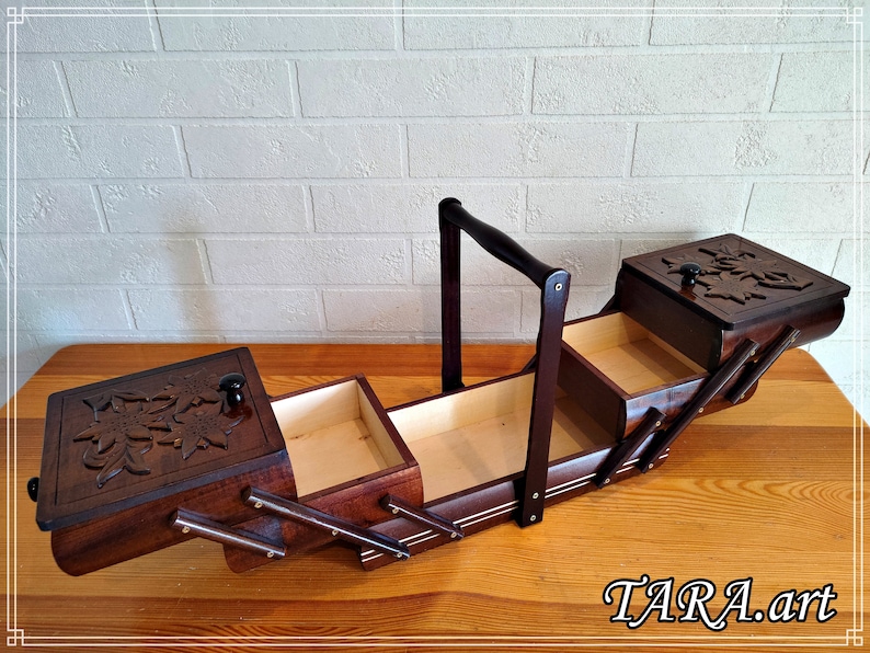 May include: A wooden sewing box with a hinged lid and a handle. The box has three compartments and is decorated with carved floral designs. The box is open and the compartments are empty.
