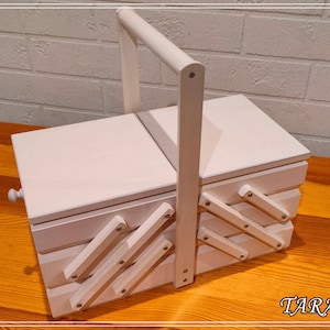 May include: A white wooden sewing box with a hinged lid and a handle. The box has a decorative design on the front and sides.