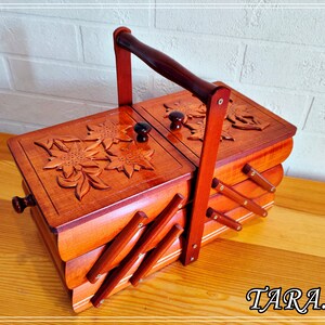 May include: A wooden sewing box with a carved floral design and a handle. The box has three compartments and is made of light brown wood.