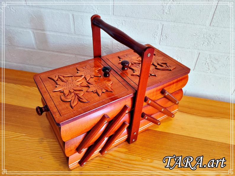 May include: A wooden sewing box with a carved floral design and a handle. The box has three compartments and is made of light brown wood.