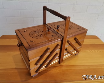 May include: A wooden sewing box with a carved design on the lid and a handle. The box has multiple compartments and is made of light brown wood.