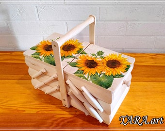 May include: A light-coloured wooden sewing box with a cantilevered accordion design and a central handle. The top lids feature a vibrant sunflower print with yellow petals and green leaves. The text TARA.art is visible in the bottom right corner.
