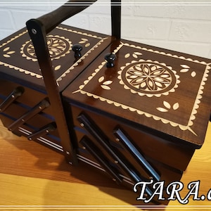 Puede incluir: Una caja de costura de madera con asa y tallas florales intrincadas. La caja tiene dos compartimentos y una tapa con bisagras. La caja es marrón con una incrustación de madera clara.