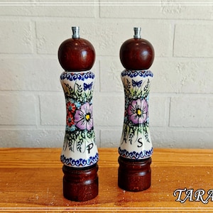 Könnte beinhalten: Zwei Salz- und Pfeffermühlen aus Holz mit einem weißen Keramikkörper, der mit einem handgemalten Blumenmuster in Lila, Rot und Grün sowie blauen Schmetterlingen verziert ist. Die Mühlen haben dunkelbraune Holzober- und -unterteile mit den Buchstaben P und S.