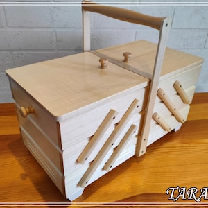 May include: A wooden sewing box with a hinged lid and a handle. The box has a decorative design on the front and sides, and it is made of light wood.