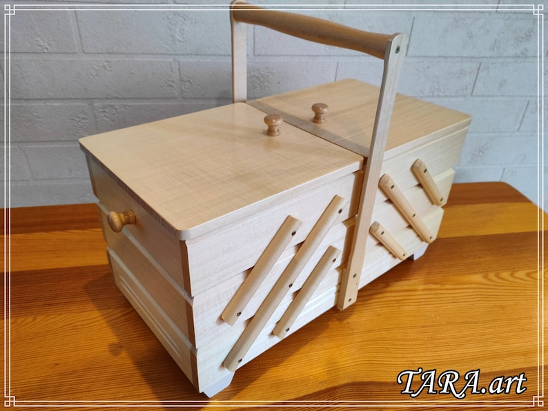 May include: A wooden sewing box with a hinged lid and a handle. The box has a decorative design on the front and sides, and it is made of light wood.