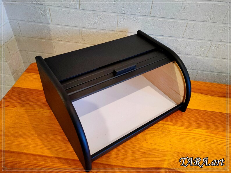 May include: Black wooden bread box with a hinged lid and a small handle. The box is sitting on a wooden surface.