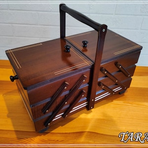 Puede incluir: Una caja de costura de madera marrón oscuro con un asa y perillas negras. La caja tiene múltiples compartimentos y cajones, con detalles dorados y blancos. La caja está sobre una superficie de madera.