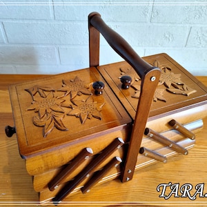Puede incluir: Una caja de costura de madera con un diseño floral tallado en la tapa. La caja tiene un asa y tres compartimentos. La caja está hecha de madera marrón claro.