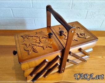 May include: A wooden sewing box with a carved floral design on the lid. The box has a handle and three compartments. The box is made of light brown wood.