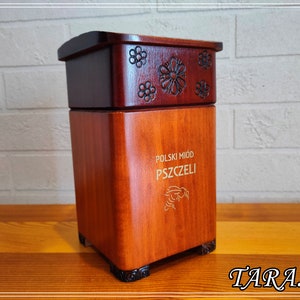 May include: A wooden honey container with a dark brown lid and a lighter brown base. The lid is decorated with carved floral designs. The base has text in Polish and a bee illustration. The container is rectangular with rounded corners.