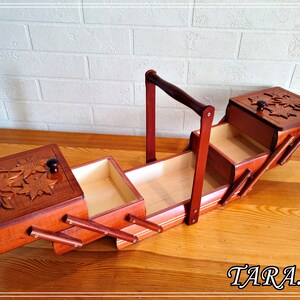 May include: A wooden sewing box with a hinged lid and a carved floral design. The box has four compartments and a handle for carrying. The box is open and the compartments are visible.
