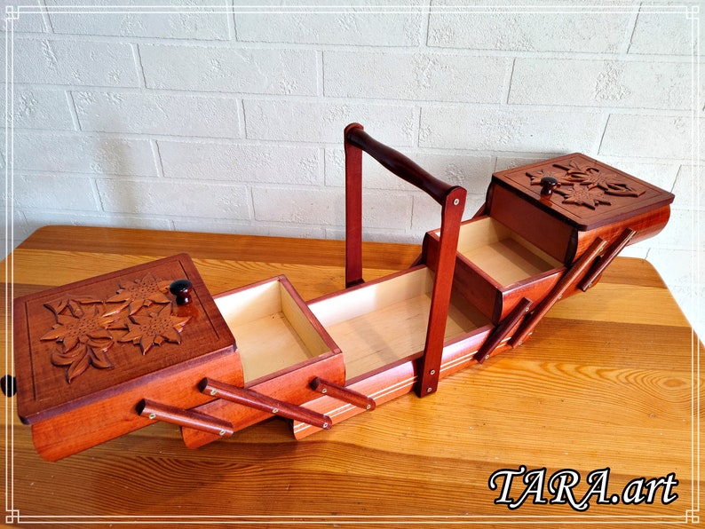 May include: A wooden sewing box with a hinged lid and a carved floral design. The box has four compartments and a handle for carrying. The box is open and the compartments are visible.