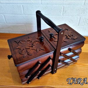 May include: A wooden sewing box with a carved floral design on the lid. The box has a handle and is divided into three sections. The box is brown and has a black trim.