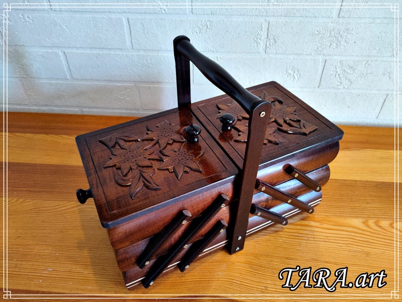 May include: A wooden sewing box with a carved floral design on the lid. The box has a handle and is divided into three sections. The box is brown and has a black trim.