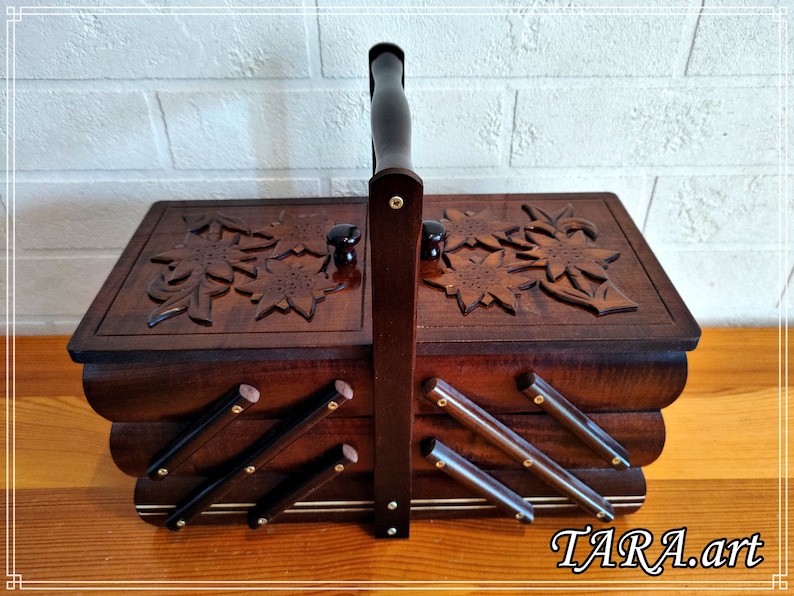 May include: A wooden sewing box with a carved floral design and a handle. The box has three compartments and is made of dark wood.