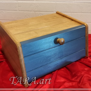 Op de afbeelding: Een houten broodtrommel met een blauw geverfde voorkant en een houten knop. De broodtrommel heeft drie horizontale groeven aan de voorkant.