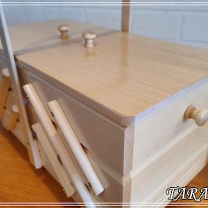 May include: A wooden sewing box with a hinged lid and a drawer. The box is made of light wood and has a natural finish. The lid has two small wooden knobs for opening and closing. The drawer has a single knob. The box is sitting on a wooden surface.