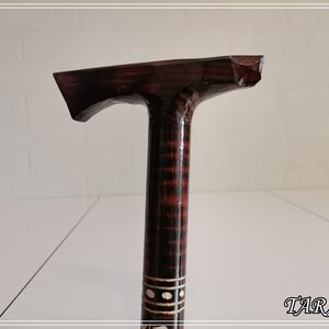May include: A wooden walking stick with a dark brown and black striped pattern. The top of the stick is shaped like a T and has a dark brown finish. The stick is decorated with a white band with black dots.