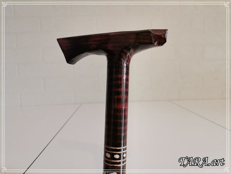 May include: A wooden walking stick with a dark brown and black striped pattern. The top of the stick is shaped like a T and has a dark brown finish. The stick is decorated with a white band with black dots.