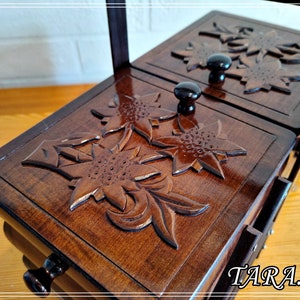 May include: A wooden box with a hinged lid, featuring intricate carved floral designs. The box is made of dark brown wood and has a polished finish. The lid is open, revealing an empty interior.