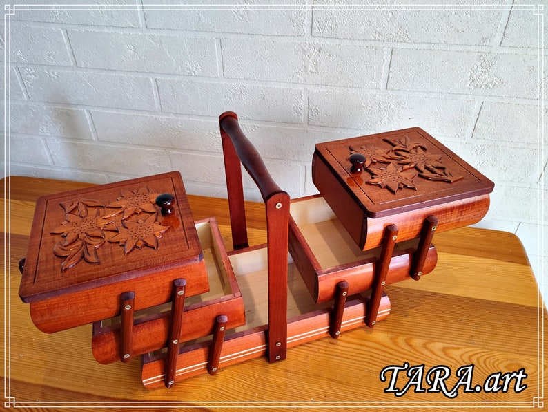 May include: A wooden sewing box with three compartments and a handle. The box features a carved floral design on the lids and is made from rich brown wood.