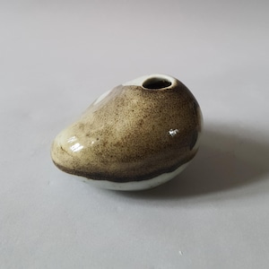 Op de afbeelding: Een kleine keramische vaas met een afgeronde, kiezelachtige vorm. De vaas heeft een gespikkelde bruine en beige glazuur over een witte basis, met een kleine opening aan de bovenkant. Het ontwerp is minimalistisch en organisch.