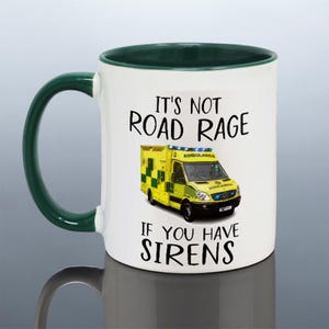 May include: White ceramic mug with a green rim. The mug features a yellow ambulance with blue lights and the text "It's not road rage if you have sirens".