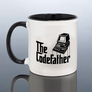 May include: A white ceramic mug with a black interior and a black handle. The exterior features a black graphic illustration of a vintage computer terminal positioned above the black text that reads The Codefather in a stylised gothic font.