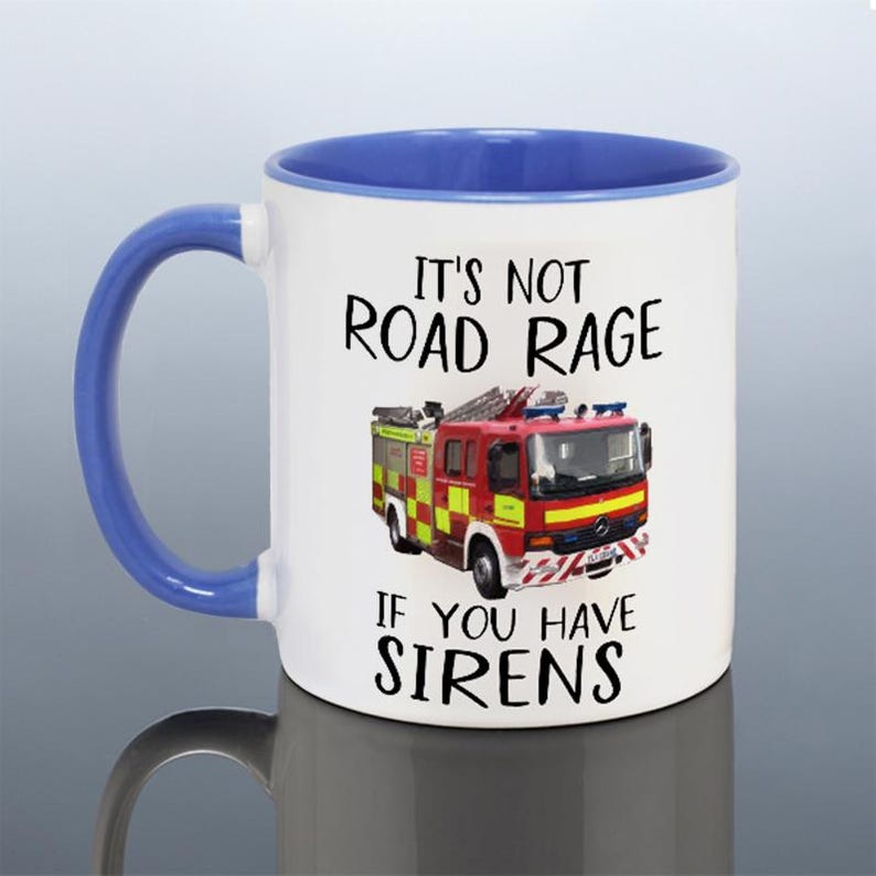 May include: White ceramic mug with a blue handle and interior. The mug features a red and yellow fire truck graphic and the text "IT'S NOT ROAD RAGE IF YOU HAVE SIRENS."
