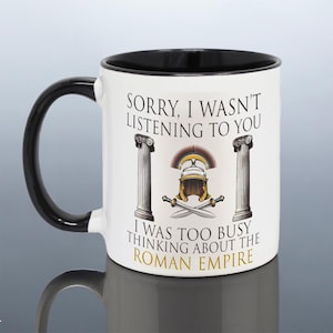 May include: A white ceramic mug with a black handle and interior rim. The front features a graphic of two grey stone columns flanking a Roman helmet and crossed swords. The text reads: SORRY, I WASN'T LISTENING TO YOU I WAS TOO BUSY THINKING ABOUT THE ROMAN EMPIRE.