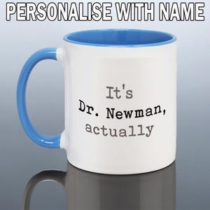 May include: A white ceramic mug with a bright blue handle and interior. The text "PERSONALISE WITH NAME" appears at the top. The front features the black printed text "It's Dr. Newman, actually" in a typewriter-style font on the white surface.