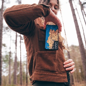 K&ouml;nnte beinhalten: Eine Person tr&auml;gt eine braune Cordjacke und eine schwarze Leggings und h&auml;lt eine blaue und goldene Handyh&uuml;lle mit einem Ausschnitt f&uuml;r das Kameraobjektiv. Die Handyh&uuml;lle hat ein einzigartiges Holzmaserungsmuster.