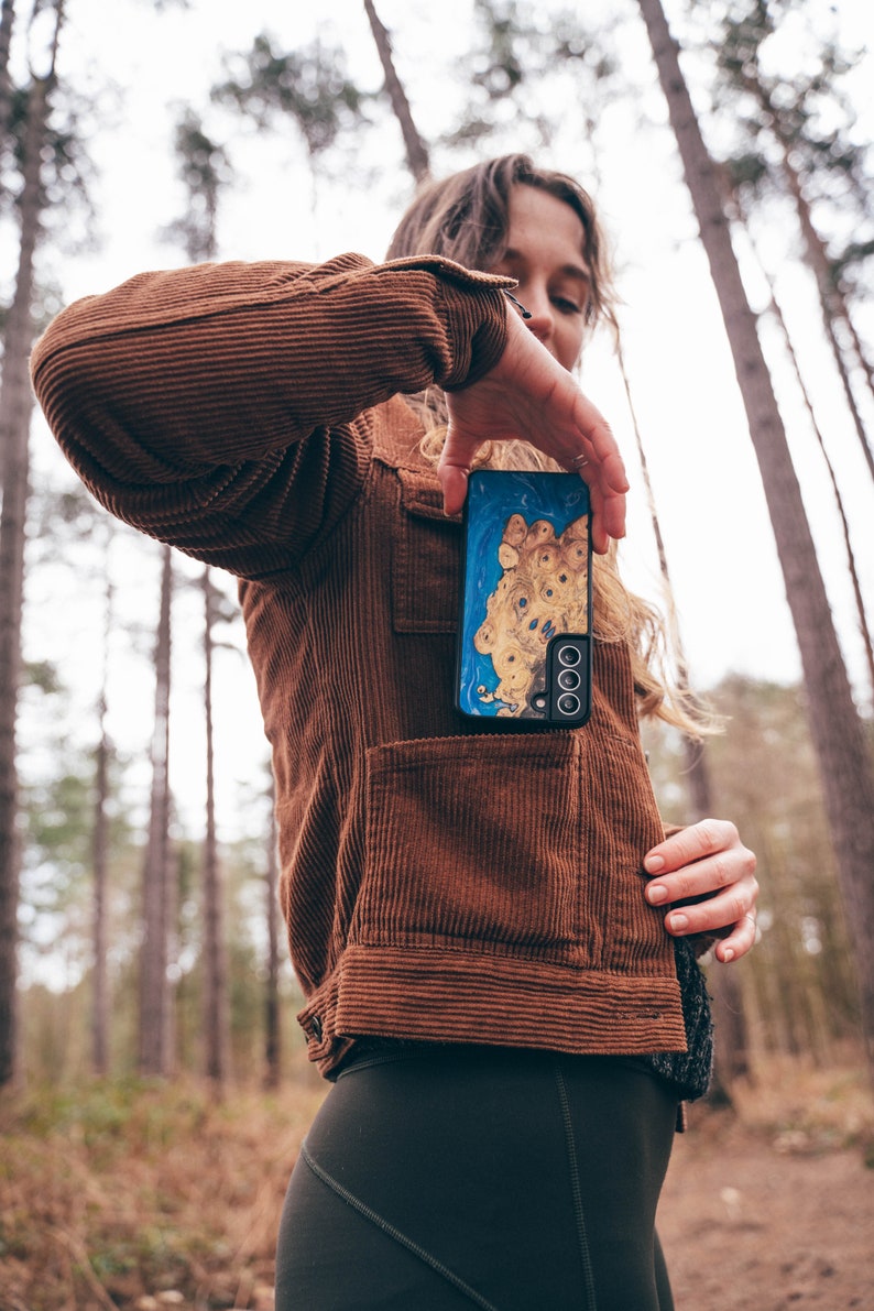 K&ouml;nnte beinhalten: Eine Person tr&auml;gt eine braune Cordjacke und eine schwarze Leggings und h&auml;lt eine blaue und goldene Handyh&uuml;lle mit einem Ausschnitt f&uuml;r das Kameraobjektiv. Die Handyh&uuml;lle hat ein einzigartiges Holzmaserungsmuster.