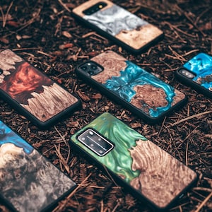 May include: A collection of phone cases with unique designs. Each case features a wooden base with colorful resin accents in shades of blue, green, red, and silver. The cases are black and rest on a bed of brown leaves and twigs.