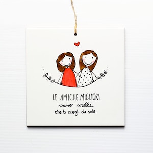 Op de afbeelding: Een witte keramische tegel met een handgetekend ontwerp van twee vriendinnen die elkaars hand vasthouden. De tekst luidt "LE AMICHE MIGLIORI sono sorelle che ti scegli da sola."