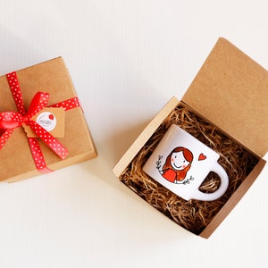 Puede incluir: Una taza de cerámica blanca con una ilustración dibujada a mano de una persona con cabello rojo y un corazón rojo. La taza está en una caja de cartón marrón con papel triturado. La caja está atada con una cinta roja con lunares blancos.