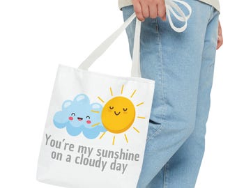 Peut inclure: Sac fourre-tout blanc avec un motif de soleil et de nuage dessinés. Le soleil et le nuage ont des visages souriants. Le texte sur le sac est "You're my sunshine on a cloudy day."